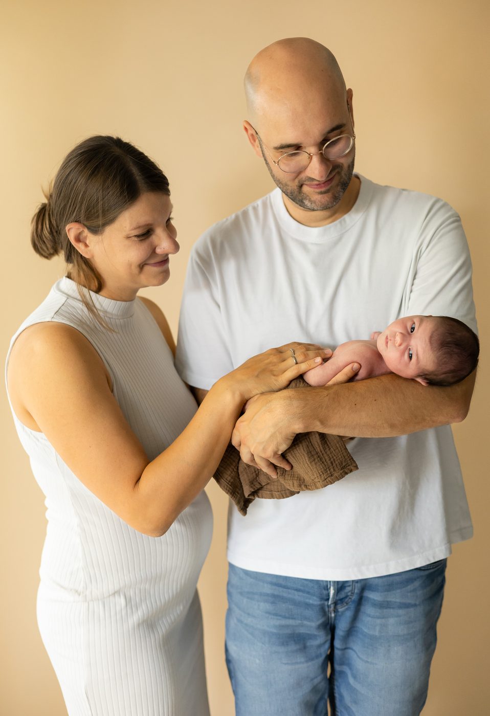Die Eltern stehen zusammen, der Papa h&auml;lt das Baby, die Mama l&auml;chelt es an